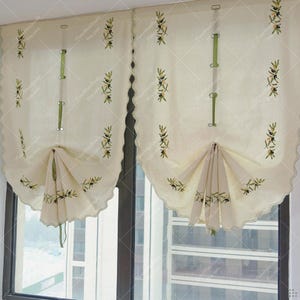 May include: Cream-colored window curtains with scalloped edges and olive branch embroidery. The curtains have a gathered design and are held up with green straps. The olive branch design features green leaves and black olives.