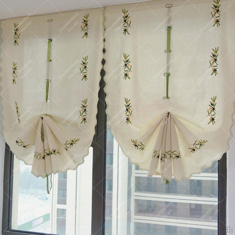 May include: Cream-colored window curtains with scalloped edges and olive branch embroidery. The curtains have a gathered design and are held up with green straps. The olive branch design features green leaves and black olives.