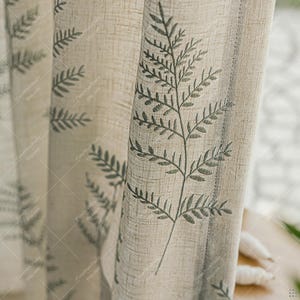 Puede incluir: Cortinas de lino beige con un patrón repetitivo de hojas de helecho en verde apagado. Las cortinas tienen una apariencia texturizada y están parcialmente corridas, revelando una ventana y luz natural.