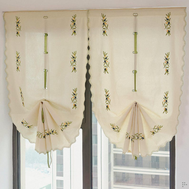 May include: Cream-colored window curtains with olive branch embroidery. The curtains have a scalloped edge and are gathered at the bottom with green ribbon ties. The design features embroidered olive branches in green and black.