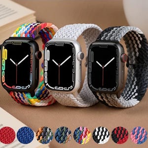 Op de afbeelding: Drie smartwatches met gevlochten bandjes in regenboog-, witte en zwart-grijze patronen. De wijzerplaten van de horloges zijn zwart met witte wijzers. Een selectie van bandkleuren, waaronder rood, blauw en zwart, wordt hieronder weergegeven.