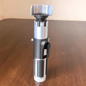 May include: A silver, black, and gray lightsaber prop. The hilt features a black grip section, a silver midsection, and a gray emitter. The lightsaber is standing upright on a wooden surface.
