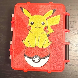 May include: Red plastic case featuring a yellow Pikachu character sitting on a Poke Ball. The case has a hinged lid with black and red accents. The design is a vibrant and colorful representation of the popular animated series.