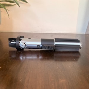 May include: A silver and black lightsaber prop, placed on a dark wooden surface. The prop has a detailed handle with ribbed and buttoned sections, and a silver blade emitter. The lightsaber is a replica of a popular science fiction weapon.