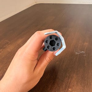 May include: A black, cylindrical object with a hexagonal center and multiple circular holes, held with a silver wrench. The object is likely a tool or part of a mechanism. The background is a wooden surface.