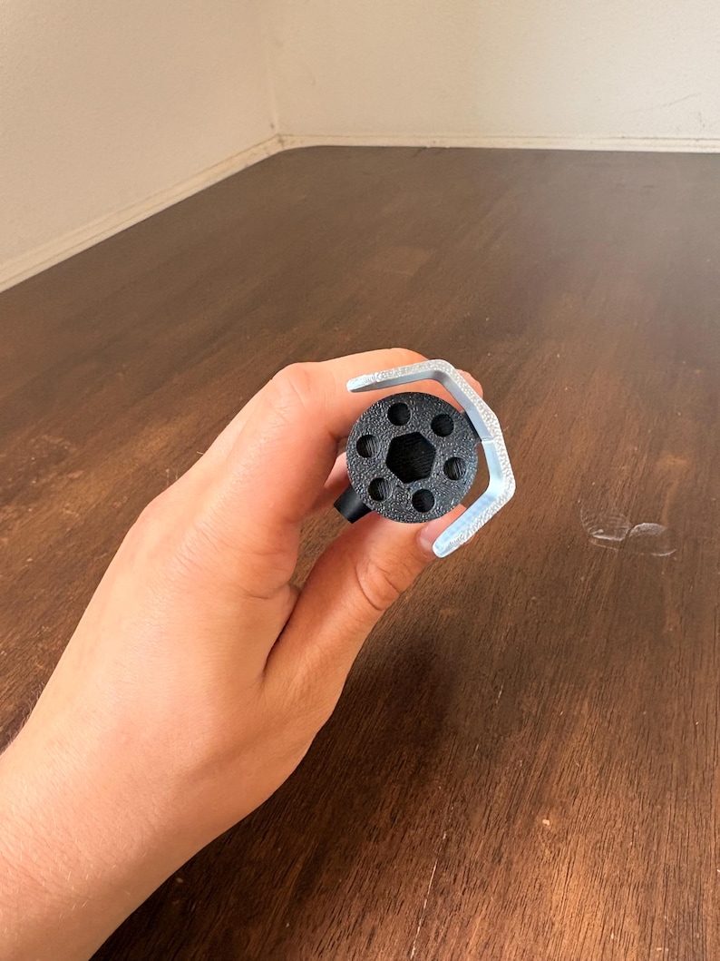 May include: A black, cylindrical object with a hexagonal center and multiple circular holes, held with a silver wrench. The object is likely a tool or part of a mechanism. The background is a wooden surface.