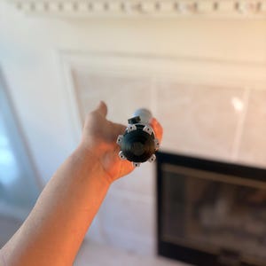 Pu&ograve; includere: Uno strumento nero e argento con una testa circolare e pi&ugrave; punte &egrave; tenuto in mano. Lo strumento &egrave; cilindrico e sembra essere in metallo. Lo sfondo &egrave; un interno sfocato con un camino.