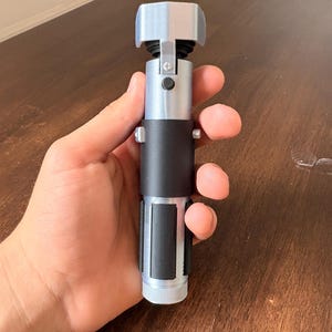 May include: A silver and black lightsaber prop with a rectangular emitter and a cylindrical handle. The handle has a black grip and a silver base. The prop is held in a hand.