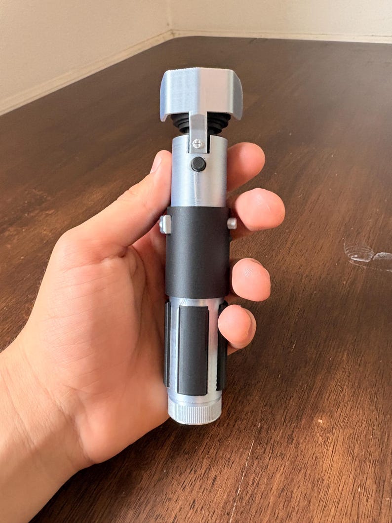 May include: A silver and black lightsaber prop with a rectangular emitter and a cylindrical handle. The handle has a black grip and a silver base. The prop is held in a hand.