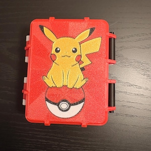 May include: A red plastic case featuring a yellow Pikachu character sitting on a Poke Ball. The case has black hinges and white clasps. The background is a dark wood surface.