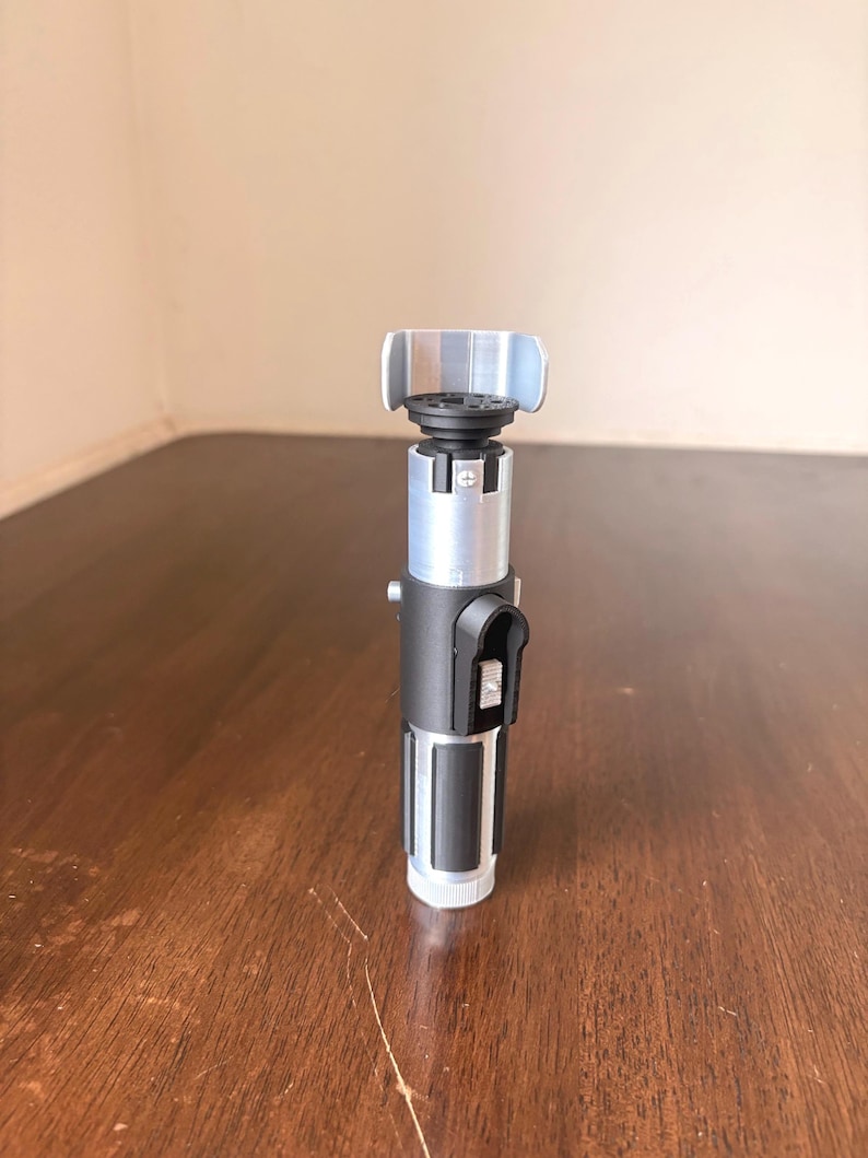 May include: A silver, black, and gray lightsaber prop. The hilt features a black grip section, a silver midsection, and a gray emitter. The lightsaber is standing upright on a wooden surface.