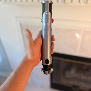 Pu&ograve; includere: Una spada laser argentata con dettagli neri e rossi. L'elsa presenta un design dettagliato con pulsanti e un'impugnatura strutturata. La spada laser &egrave; tenuta in mano, mostrando le sue dimensioni e il suo design.