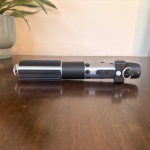 May include: A black and silver lightsaber prop with a cylindrical design. The hilt features a ribbed grip and a silver finish with black accents. The lightsaber has a ring and a button on the end.