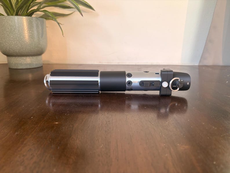 May include: A black and silver lightsaber prop with a cylindrical design. The hilt features a ribbed grip and a silver finish with black accents. The lightsaber has a ring and a button on the end.