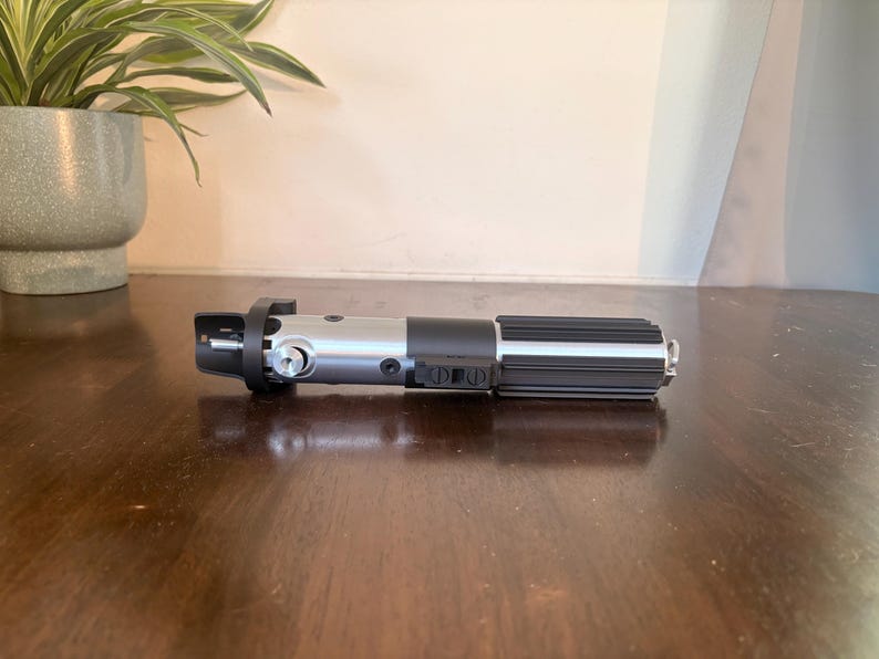 May include: A silver and black lightsaber prop, placed on a dark wooden surface. The prop has a detailed handle with ribbed and buttoned sections, and a silver blade emitter. The lightsaber is a replica of a popular science fiction weapon.
