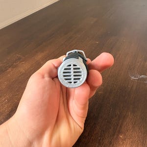 May include: A silver-colored, cylindrical object with a series of horizontal slits, held in a hand. The object has a black strap and a small, rounded top. The background is a wooden surface.