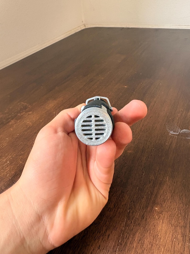 May include: A silver-colored, cylindrical object with a series of horizontal slits, held in a hand. The object has a black strap and a small, rounded top. The background is a wooden surface.