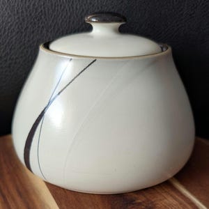 以下が含まれることがあります： クリーム色のセラミックシュガーボウルと蓋。ボウルは丸みを帯びた形状で、濃い茶色のアクセントラインと、お揃いの蓋のハンドルが付いています。ボウルは木製の表面に置かれています。