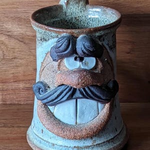 Può includere: Una tazza in ceramica fatta a mano con un design a faccina sorridente. La tazza ha una base azzurra, un bordo marrone e un manico. Il viso ha baffi marroni, sopracciglia e un ampio sorriso con denti bianchi.