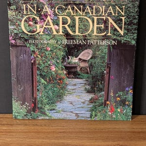 May include: A book cover with the title "In a Canadian Garden" by Nicole Eaton & Hilary Weston. The cover features a photograph of a garden path leading to a wicker chair, with lush greenery and flowers.
