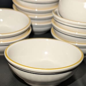 Peut inclure: Ensemble de bols en céramique blanche, bordés d'un liseré jaune et d'un trait noir. Les bols sont empilés, présentant un design simple et classique. Idéal pour servir soupes, céréales ou collations.