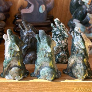 May include: A collection of carved stone wolf figurines in various shades of green, gray, and white. The wolves are depicted howling with open mouths and are displayed on a wooden shelf. The figurines vary in size and texture.