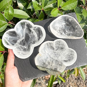 May include: Three clear, polished quartz crystal bowls with smooth, rounded edges. Two of the bowls feature a distinct four-lobed, flower-like shape, while the third bowl is a smaller, irregular triangular form. All items are transparent.