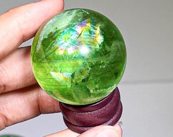 May include: A polished, translucent green sphere with internal fractures and a vibrant rainbow-colored light reflection. The sphere is securely seated on a small, dark reddish-brown wooden stand, showcasing its smooth, rounded surface and depth.