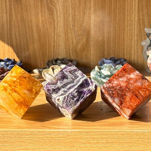 May include: Three gemstone cubes in shades of yellow, purple, and red-orange. The yellow cube has a layered appearance, the purple cube has white veining, and the red-orange cube has a speckled pattern. The cubes are displayed on a wooden surface.
