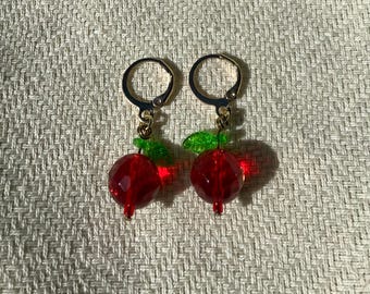 May include: A pair of gold-toned hoop earrings, each featuring a faceted red bead resembling a cherry. Small green leaf accents and a gold-toned stem complete the cherry design. The earrings are displayed against a textured, off-white background.