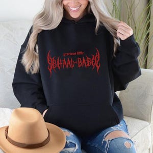 Könnte beinhalten: Schwarzer Kapuzenpullover mit dem Text "precious little SBHAAL BABE" in roter gotischer Schrift. Der Pullover hat eine Vordertasche und eine Kapuze mit Kordelzug. Ein beigefarbener Cowboyhut mit braunem Lederband ist im Vordergrund.