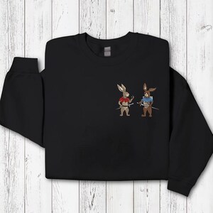 Puede incluir: Sudadera negra con conejos de dibujos animados bordados vestidos de guerreros. Un conejo lleva una camiseta roja, el otro una camiseta azul, ambos sosteniendo espadas. El dise&ntilde;o est&aacute; centrado en el pecho.