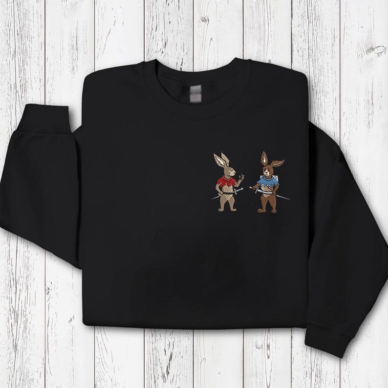 Puede incluir: Sudadera negra con conejos de dibujos animados bordados vestidos de guerreros. Un conejo lleva una camiseta roja, el otro una camiseta azul, ambos sosteniendo espadas. El dise&ntilde;o est&aacute; centrado en el pecho.