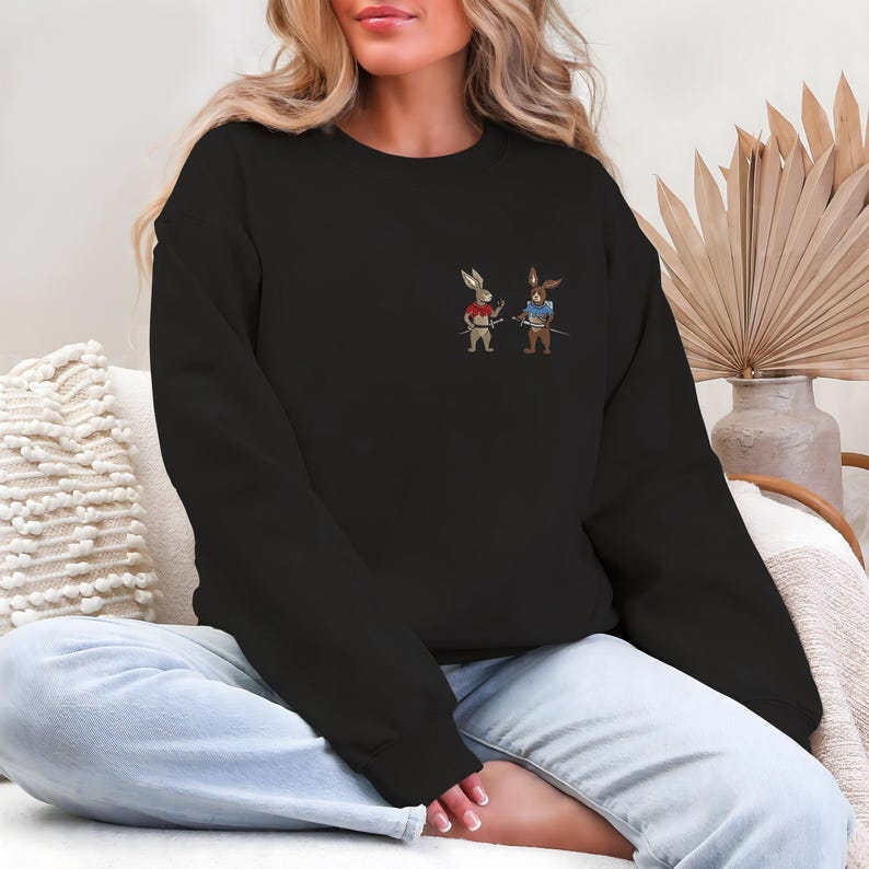 Puede incluir: Sudadera negra con un dise&ntilde;o bordado de dos conejos de dibujos animados vestidos de samur&aacute;is, cada uno sosteniendo una espada. Un conejo viste de rojo, el otro de azul. La sudadera es de estilo casual.
