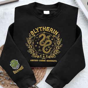 May include: Black sweatshirt featuring a gold embroidered "Slytherin" design, a snake, and the words "Ambitious, Cunning, Resourceful." The left sleeve has a green and gold patch with a snake and the word "Greatness."
