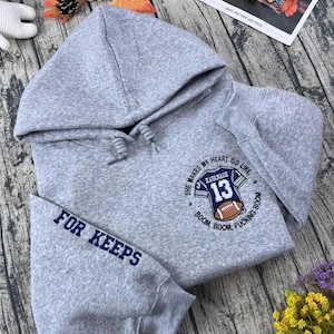 May include: A gray hooded sweatshirt with the text "FOR KEEPS" on the sleeve. The front features a graphic of a football jersey with the number 13 and the words "SHE MAKES MY HEART GO LIKE" and "BOOM, BOOM, FUCKING BOOM."