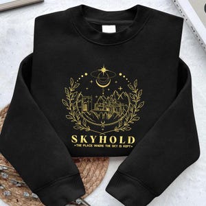 Op de afbeelding: Zwart sweatshirt met een gouden grafisch ontwerp met een kasteel, bergen en hemelse elementen. De tekst "SKYHOLD" en "THE PLACE WHERE THE SKY IS KEPT" staan eronder. Het sweatshirt is op een geweven oppervlak gevouwen.