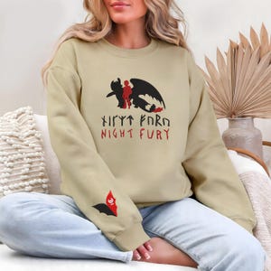 Könnte beinhalten: Beiges Sweatshirt mit einem schwarzen und roten Drachen-Motiv und dem Text "Night Fury". Das Design beinhaltet eine Drachensilhouette und eine Figur. Auf dem Ärmel befindet sich eine kleinere fledermausartige Grafik.