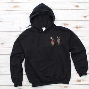 Puede incluir: Sudadera con capucha negra con bolsillo de canguro. La sudadera presenta un dise&ntilde;o bordado de dos conejos vestidos de samur&aacute;is. Uno de los conejos lleva un chaleco rojo, el otro un chaleco azul, ambos con espadas.