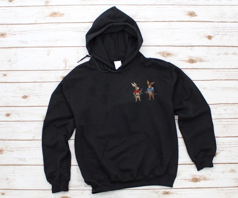 Puede incluir: Sudadera con capucha negra con bolsillo de canguro. La sudadera presenta un dise&ntilde;o bordado de dos conejos vestidos de samur&aacute;is. Uno de los conejos lleva un chaleco rojo, el otro un chaleco azul, ambos con espadas.