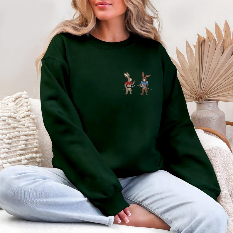 Puede incluir: Sudadera verde oscuro con un dise&ntilde;o bordado de dos conejos samur&aacute;is. Uno es rojo, el otro azul, ambos con espadas. La sudadera es de color liso y tiene cuello redondo.