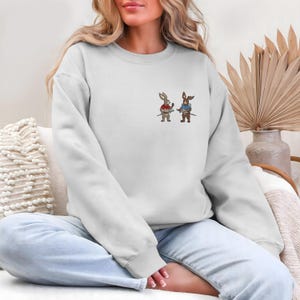 Puede incluir: Una sudadera blanca con un dise&ntilde;o bordado de dos conejos de dibujos animados sosteniendo espadas. Un conejo lleva una camisa roja, el otro una camisa azul. La sudadera tiene mangas largas y cuello redondo.