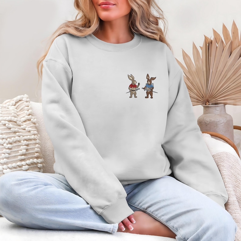 Puede incluir: Una sudadera blanca con un dise&ntilde;o bordado de dos conejos de dibujos animados sosteniendo espadas. Un conejo lleva una camisa roja, el otro una camisa azul. La sudadera tiene mangas largas y cuello redondo.