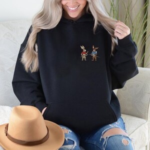 Puede incluir: Sudadera con capucha negra con bolsillo de canguro. La sudadera presenta un peque&ntilde;o dise&ntilde;o bordado de dos conejos de dibujos animados vestidos de samur&aacute;is. Los conejos sostienen espadas. Un sombrero de vaquero marr&oacute;n est&aacute; en primer plano.