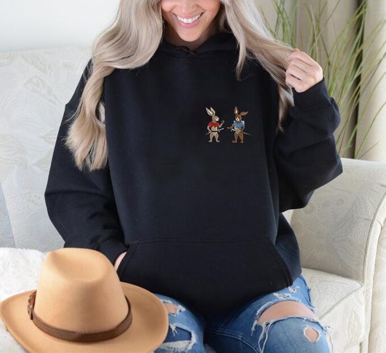 Puede incluir: Sudadera con capucha negra con bolsillo de canguro. La sudadera presenta un peque&ntilde;o dise&ntilde;o bordado de dos conejos de dibujos animados vestidos de samur&aacute;is. Los conejos sostienen espadas. Un sombrero de vaquero marr&oacute;n est&aacute; en primer plano.
