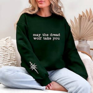 May include: A dark green sweatshirt with the text "may the dread wolf take you" in white. A white graphic of a sword and sunburst design is on the left sleeve. The sweatshirt is a casual garment.