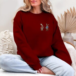 Puede incluir: Una sudadera burdeos con un peque&ntilde;o dise&ntilde;o bordado de dos conejos de dibujos animados vestidos de guerreros. Un conejo viste de rojo, el otro de azul, cada uno con una espada. La sudadera es de color liso y tiene cuello redondo.