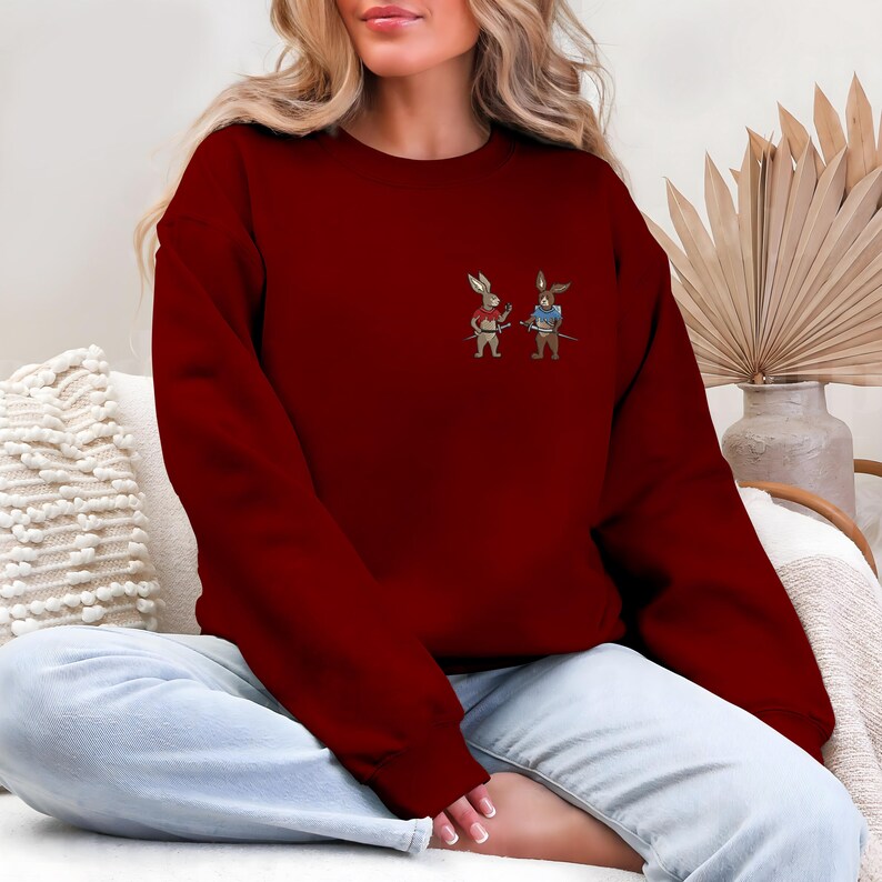 Puede incluir: Una sudadera burdeos con un peque&ntilde;o dise&ntilde;o bordado de dos conejos de dibujos animados vestidos de guerreros. Un conejo viste de rojo, el otro de azul, cada uno con una espada. La sudadera es de color liso y tiene cuello redondo.