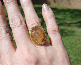 May include: A gold ring featuring a large, oval-shaped rutilated quartz gemstone. The stone is translucent with visible golden needle-like inclusions throughout the interior. The setting is a smooth, polished gold band that encircles the gemstone.