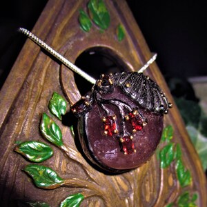 May include: A pendant necklace with a dark purple gemstone and red accents, featuring a dark metal frog design. The pendant hangs from a silver chain. The pendant is set against a wooden tree-like structure with green leaves.